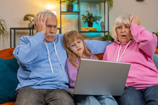 Upset senior grandfather grandmother and granddaughter playing game on laptop and losing at home. Sad Caucasian girl with grandparents received bad news fortune loss fail education study test on sofa