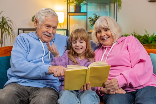 Caucasian grandparents and little granddaughter reading interesting book together sitting on sofa in living room at home. Girl with grandparents spending leisure time on comfortable couch in apartment