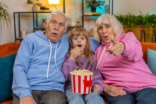 Amazed Caucasian grandfather grandmother and granddaughter eating popcorn and watching movie on sofa at home. Smiling small girl with grandparents enjoying film during weekend in living room apartment