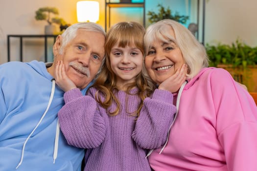 Portrait of smiling kid granddaughter and loving senior grandparents looking at camera. Girl child with grandfather and grandmother embracing sitting on sofa in living room at home. Love bonding care.