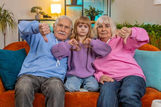 Portrait of upset grandparents and granddaughter showing thumbs down gesture, expressing discontent, disapproval, dissatisfied. Displeased family looking at camera on couch in living room at home