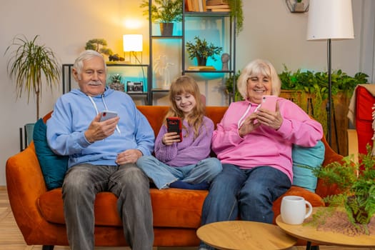 Addicted grandfather, grandmother and granddaughter using smartphones in living room at home. Smiling girl and senior couple spending leisure time sitting on sofa. Playing online games, social media.