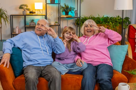 Happy excited Caucasian senior grandparents and granddaughter girl dancing together in disco style on home sofa. Smiling family enjoying weekends together in apartment. Carefree fun freedom concepts