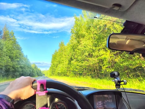 Car salon, windshield, hand of woman on steering wheel and landscape. View from seat of driver on nature with Road, trees, blue sky at a sunny day. Single trip of female traveler. Partial focus