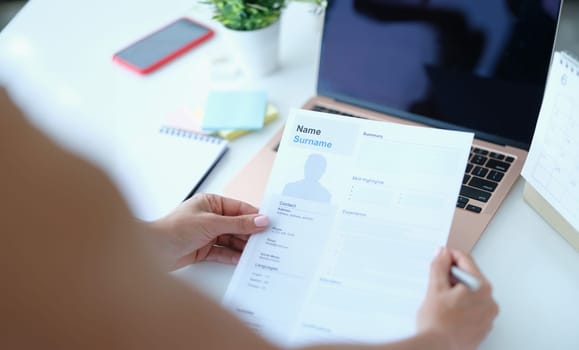 Woman sitting at table in front of laptop and holding paper with resume for employment in her hands. Online interview when applying for job concept
