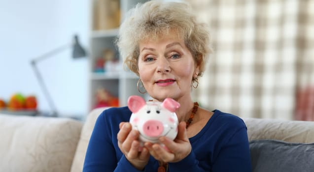 Happy elderly woman shows pink ceramic piggy bank. Initial savings fund. Expenses include food. Successful budgeting. Accumulation and economy. Control and analysis income and expenses