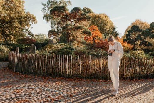 Beautiful young woman takes photos with a professional camera in autumn forest. Smiling girl enjoying autumn weather. Rest, relaxation, lifestyle concept. Young photographer takes pictures of autumn forest at sunset