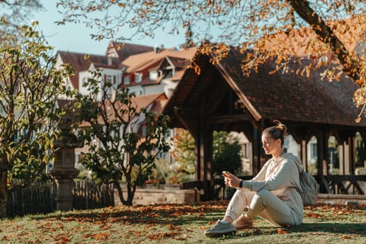 Young fashionable teenage girl with smartphone in park in autumn sitting at smiling. Trendy young woman in fall in park texting. Retouched, vibrant colors. Beautiful blonde teenage girl wearing casual modern autumn outfit sitting in park in autumn. Retouched, vibrant colors, brownish tones.