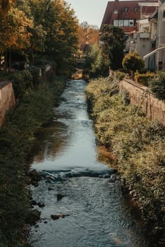 Beautiful small river with clean and clear water front of colorful autumn trees and small old town on the hill agaist nice blue and clouds sky during autumn in Europe.