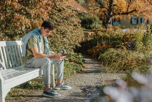 a teenager sits on a bench in the park drinks coffee from a thermo mug and looks into a phone. Portrait of handsome cheerful guy sitting on bench fresh air using device browsing media smm drinking latte urban outside outdoor.