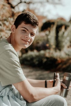 a teenager sits on a bench in the park drinks coffee from a thermo mug and looks into a phone. Portrait of handsome cheerful guy sitting on bench fresh air using device browsing media smm drinking latte urban outside outdoor.