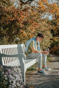 a teenager sits on a bench in the park drinks coffee from a thermo mug and looks into a phone. Portrait of handsome cheerful guy sitting on bench fresh air using device browsing media smm drinking latte urban outside outdoor.