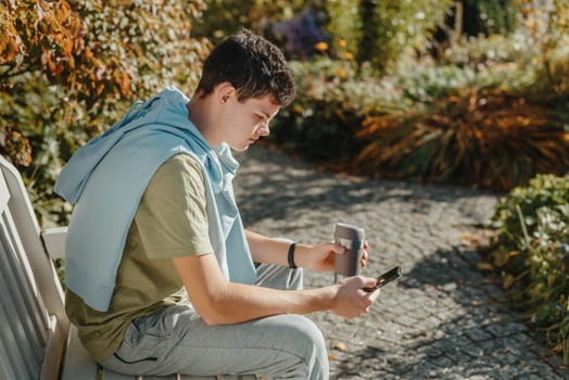 a teenager sits on a bench in the park drinks coffee from a thermo mug and looks into a phone. Portrait of handsome cheerful guy sitting on bench fresh air using device browsing media smm drinking latte urban outside outdoor.