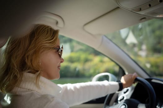 A blonde woman in a white sweater and jeans is driving. Happy woman sitting in a car with a white interior