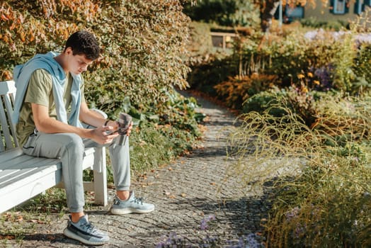 a teenager sits on a bench in the park drinks coffee from a thermo mug and looks into a phone. Portrait of handsome cheerful guy sitting on bench fresh air using device browsing media smm drinking latte urban outside outdoor.