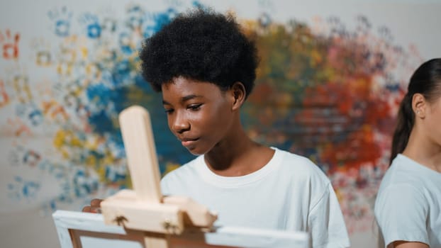 African boy painting canvas with watercolor while happy caucasian girl draw cool tone picture at colorful stained wall. Multicultural highschool student attend creative activity together. Edification.