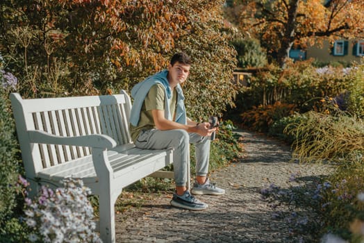 a teenager sits on a bench in the park drinks coffee from a thermo mug and looks into a phone. Portrait of handsome cheerful guy sitting on bench fresh air using device browsing media smm drinking latte urban outside outdoor.