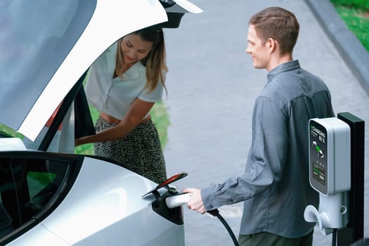 Young couple travel with EV electric car charging in green sustainable city outdoor garden in summer shows urban sustainability lifestyle by green clean rechargeable energy of electric vehicle innards