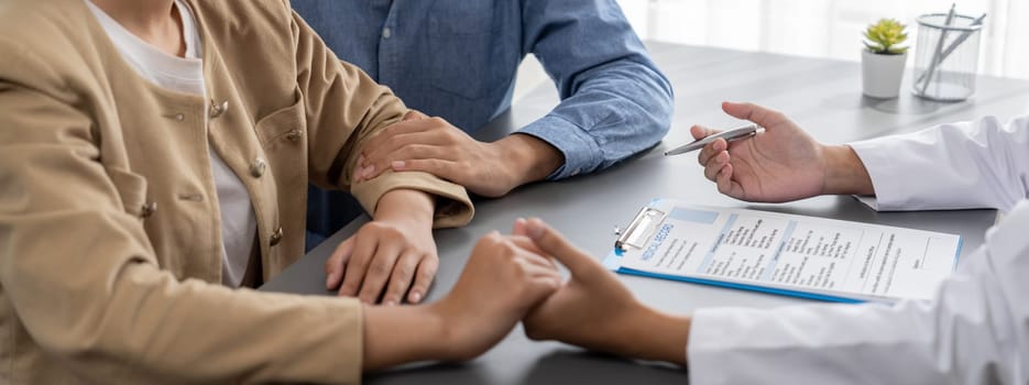 Doctor providing compassionate healthcare consultation while young couple patient holding hand, comfort each other after infertile report. Reproductive and medical fertility consulting. Neoteric