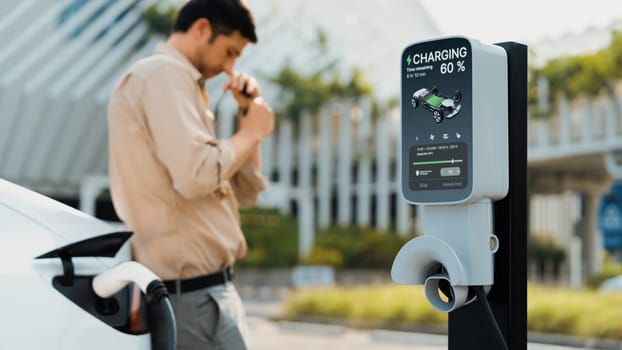 Young man put EV charger to recharge electric car's battery from charging station in city commercial parking lot. Rechargeable EV car for sustainable environmental friendly urban travel. Expedient