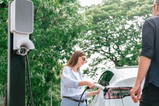 Lovely young couple wearing sun glasses recharging battery for electric car during road trip travel EV car in natural forest or national park. Eco friendly travel during vacation and holiday. Exalt