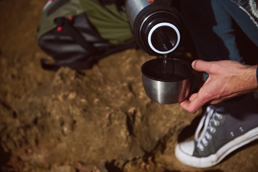 Close-up of the hands of a male tourist holding a stainless steel thermos bottle, pouring some hot drink into a cup while relaxing during his adventure trip. People. Active lifestyle. Tourism