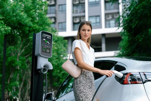 Young woman travel with EV electric car charging in green sustainable city outdoor garden in summer. Urban sustainability lifestyle by green clean rechargeable energy of electric BEV vehicle innards