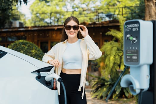 Young woman and sustainable urban commute with EV electric car recharging at outdoor cafe in springtime garden, green city sustainability and environmental friendly EV car. Expedient