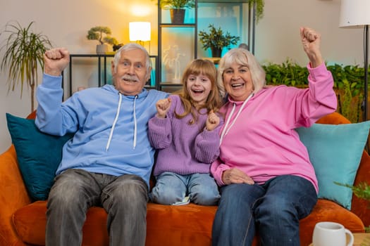 Happy Caucasian grandparents and granddaughter watching TV shouting, celebrating success, winning, goal achievement good news, victory sitting on sofa in living room. Family clenching fists at home.