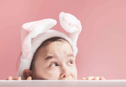 Beautiful cute caucasian girl peeking and looking to the side with headband pink and white easter bunny ears on white and pink background with little copy space ,side view closeup.Concept,banners,blanks,holiday,happy easter,childrens.