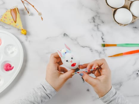 Hands of a caucasian girl in a gray turtleneck holding an easter bunny egg and gluing a colored sticker while sitting at a marble table for diy preparation for the easter holiday, flat lay closeup. The concept of crafts, needlework, art crafts, children art, handicraft, diy, artisanal.