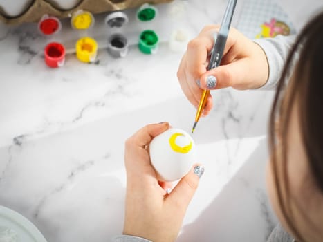 Little caucasian girl paints an easter egg with a brush with yellow acrylic paint, sitting at a marble table with a palette, willow branches, eggs in a box for diy preparation for the easter holiday, flat lay close-up. The concept of crafts, needlework, at home, children art, children creative.