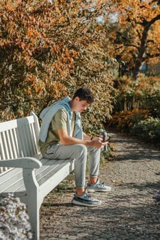 a teenager sits on a bench in the park drinks coffee from a thermo mug and looks into a phone. Portrait of handsome cheerful guy sitting on bench fresh air using device browsing media smm drinking latte urban outside outdoor.