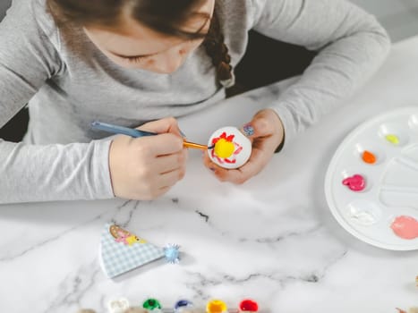 Little caucasian girl in a gray turtleneck enthusiastically paints an Easter egg with a brush of red-yellow acrylic paint at a marble table with a palette, willow branches, eggs in a box for diy preparation for the Easter holiday, close-up top view. The concept of crafts, needlework, at home,diy,artisanal,children art,children creative.
