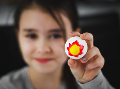 Little caucasian girl in a gray turtleneck shows an Easter egg with a red-yellow acrylic paint pattern at the table on a dark background with depth of field, close-up top view. The concept of crafts, needlework, at home, diy,children art,artisanal,preparing easter,children creative.