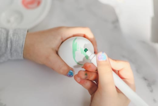 The hands of a caucasian girl in a gray turtleneck draw with a green marker on a white egg, sit at a marble table with a palette and acrylic paints, close-up side view. The concept of crafts, needlework, children's creativity, preparation for easter, children's creativity, home, crafts.