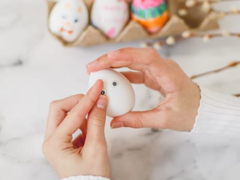 Children's hands hold one white egg and glue eye stickers, sit at a marble table with depth of field, prepare crafts for the Easter holiday, close-up side view. The concept of craft, needlework, at home, children creative, diy, crafts, children art.