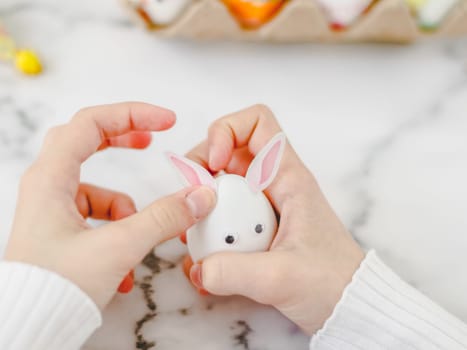 Children's hands hold one white egg with glued eyes and stick bunny ears stickers, sit at a marble table with depth of field, prepare crafts for the easter holiday, close-up side view. The concept of craft, needlework, at home, children creativity, handicraft, diy, children creative.