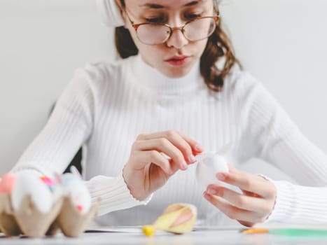 Beautiful caucasian teenage girl in a white turtleneck with glasses enthusiastically pasting stickers with bunny ears, sitting at a marble table with depth of field, preparing crafts for the Easter holiday, close-up side view. The concept of craft, needlework, at home, diy,children art,artisanal,children creative,step by step.