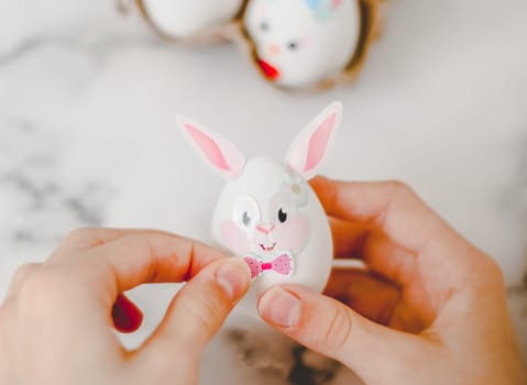 Hands of a caucasian girl pasting bow stickers on an easter bunny egg with ears, sitting at a marble table with depth of field, preparing crafts for the easter holiday, close-up side view. The concept of craft, needlework, at home, diy,children art,artisanal,children creative,step by step.