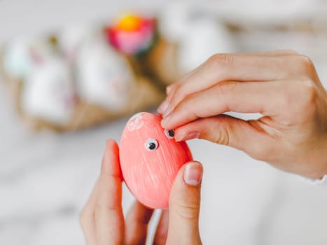 Hands of a caucasian girl sticking eye stickers on a pink acrylic painted easter egg, sitting at a marble table with depth of field, preparing crafts for the easter holiday, close-up side view. The concept of craft, needlework, at home, diy,children art,artisanal,children creative,step by step.