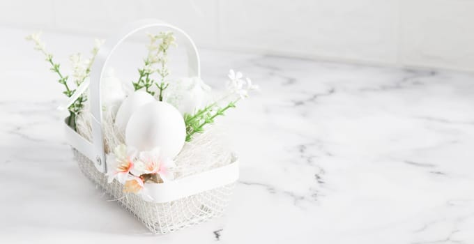 Decorative white eggs in a metal basket with eggs, spring flowers and straw stand on the left on a marble table with copy space on the right in the early morning, close-up side view. Happy easter concept, holiday banner.