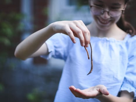 One beautiful Caucasian girl with a smile on her face holds an earthworm in her hands, standing in the garden at the backyard of a house on a spring morning, close-up side view with depth of field.