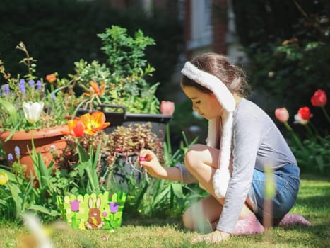 Caucasian little girl in shorts, t-shirt with sleeves, headband with bunny ears collects scattered Easter chocolate eggs in the grass while sitting on her knees and putting them in a felt basket on the lawn in the courtyard of the house, side view close up. Concept for easter tradition, culture, home, europe, picnic, easter celebration.