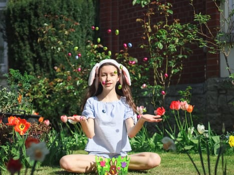 Caucasian beautiful teenage girl with headband bunny ears tosses up a chocolate easter egg while sitting on the lawn in the backyard of the house, close-up side view. Easter tradition concept, Easter celebration.