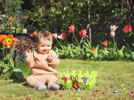 Little caucasian girl with a happy smile holds a chocolate easter egg in her hand while sitting on the lawn next to a basket and scattered candy eggs on a sunny day, close-up side view.