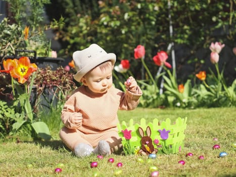 Little caucasian girl in a straw hat puts a chocolate easter egg in a felt basket while sitting on the lawn next to scattered candy eggs on a sunny day, close-up side view.