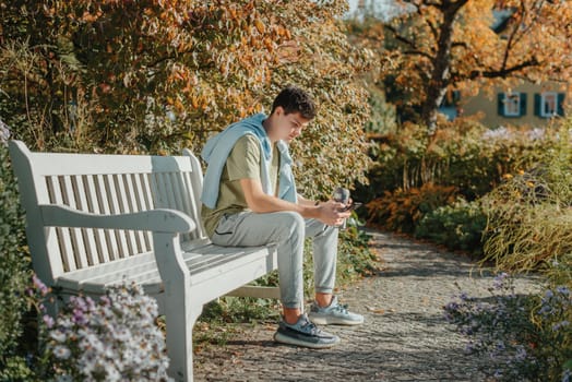 a teenager sits on a bench in the park drinks coffee from a thermo mug and looks into a phone. Portrait of handsome cheerful guy sitting on bench fresh air using device browsing media smm drinking latte urban outside outdoor.