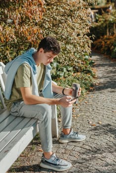 a teenager sits on a bench in the park drinks coffee from a thermo mug and looks into a phone. Portrait of handsome cheerful guy sitting on bench fresh air using device browsing media smm drinking latte urban outside outdoor.