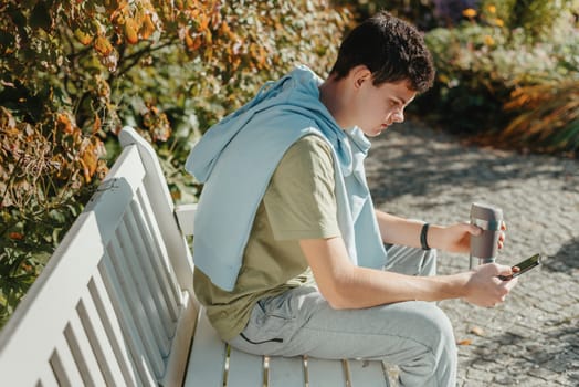 a teenager sits on a bench in the park drinks coffee from a thermo mug and looks into a phone. Portrait of handsome cheerful guy sitting on bench fresh air using device browsing media smm drinking latte urban outside outdoor.